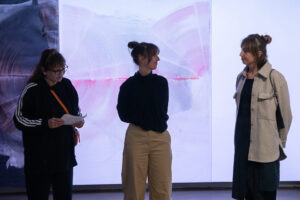 Foto eines Ausstellungsbereiches mit abstrakter Malerei und einer Videoprojektion. Im Vordergrund sind Menschen zu erkennen.