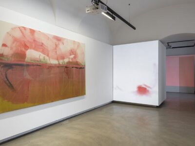 Foto eines Ausstellungsraum mit abstrakter Malerei. Man erkennt zwei Werke. Das hintere Werk dient als Projektionsleinwand.