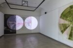 Foto eines Ausstellungsraumes mit zwei abstrakten Malereien. Die Malereien zeigen Halbkreise. Die Malerei an der Stirnwand wird von einer Projektion überlagert.