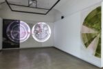 Foto eines Ausstellungsraumes mit zwei abstrakten Malereien. Die Malereien zeigen Halbkreise. Die Malerei an der Stirnwand wird von einer Projektion überlagert.
