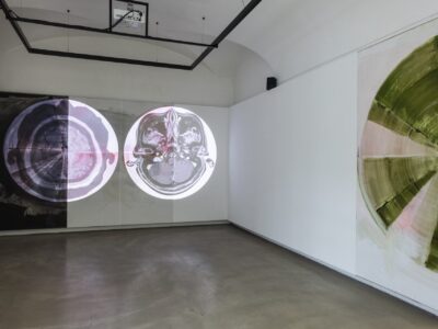 Foto eines Ausstellungsraumes mit zwei abstrakten Malereien. Die Malereien zeigen Halbkreise. Die Malerei an der Stirnwand wird von einer Projektion überlagert.