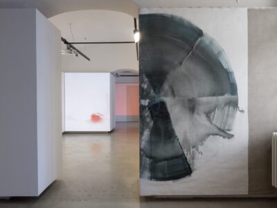 Foto eines Ausstellungsraumes mit abstrakter Malerei. Die Malereien zeigen Halbkreise. Im Hintergrund sind zwei weitere Ausstellungsräume mit abstrakter Malerei und Videoprojektion zu erkennen.