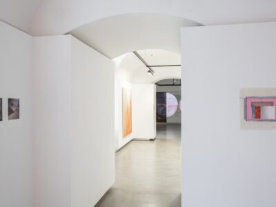 Foto eines Ausstellungsraum mit abstrakter Malerei.. Im Hintergrund erkennt man einen weiteren Ausstellungsraum mit einer Malerei an der Stirnwand, die von einer Projektion überlagert wird.