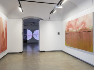 Foto eines Ausstellungsraumes mit zwei abstrakten Malereien. Die Malereien zeigen Halbkreise. Die Malerei an der Stirnwand wird von einer Projektion überlagert.