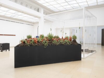 Ausstellungsraum mit schwarzem dreieckigem Beet mit Tonfiguren und Rosmarinpflanzen auf Kohle und Erdebeet