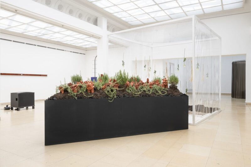 Photo of exhibition space with black objekt and on top terracotta figures and rosmary on earth and charcoal