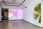 Foto eines Ausstellungsraumes mit zwei abstrakten Malereien. Die Malereien zeigen Halbkreise. Die Malerei an der Stirnwand wird von einer Projektion überlagert.