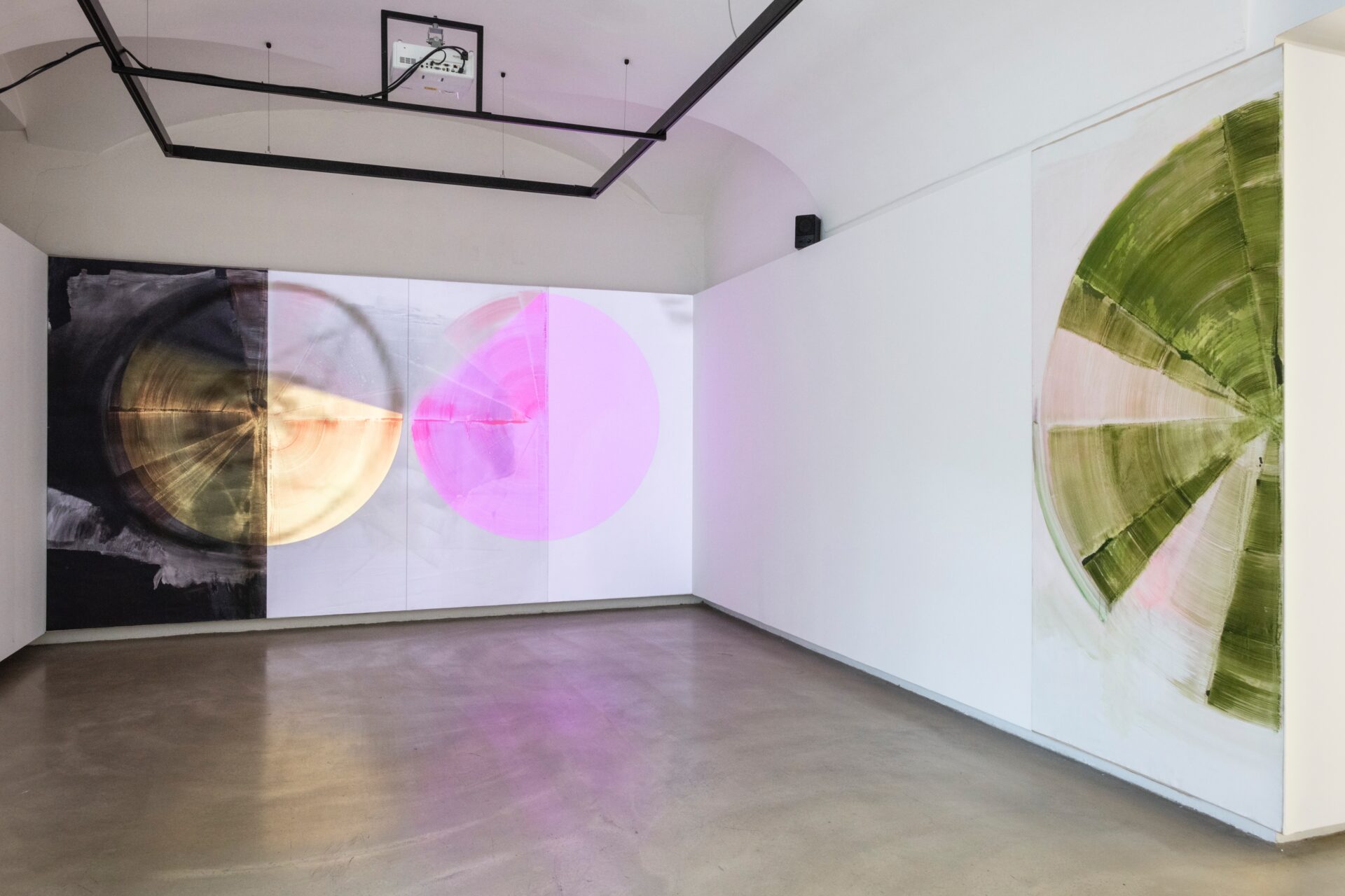 Foto eines Ausstellungsraumes mit zwei abstrakten Malereien. Die Malereien zeigen Halbkreise. Die Malerei an der Stirnwand wird von einer Projektion überlagert.