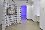 Ausstellungsansicht, im vorderen Raum sieht man einen Vorhang der in einer runden Form hängt mit Mündern darauf und ein Screen mit einem Gesicht, im hinteren Raum scheint blaues Licht