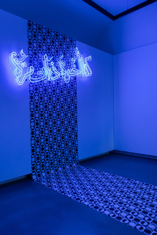 blue neon light in arabic letters saying "Once we were brothers" in front of a modern grafic wallpaper