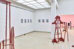 Ausstellungsansicht Kunstpavillon mit rot lackierten Holzskulpturen in unterschiedlichen Größen und Höhen mit Figurenobjekt und links sieben gemalte Bilder und ein kleines Kochtopfobjekt am Boden