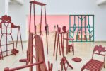 Ausstellungsansicht Kunstpavillon mit rot lackierten Holzskulpturen in unterschiedlichen Größen und Höhen und im Hintergrund ein Raumteiler mit dahinterliegenden Skulpturen vor rosa Wand