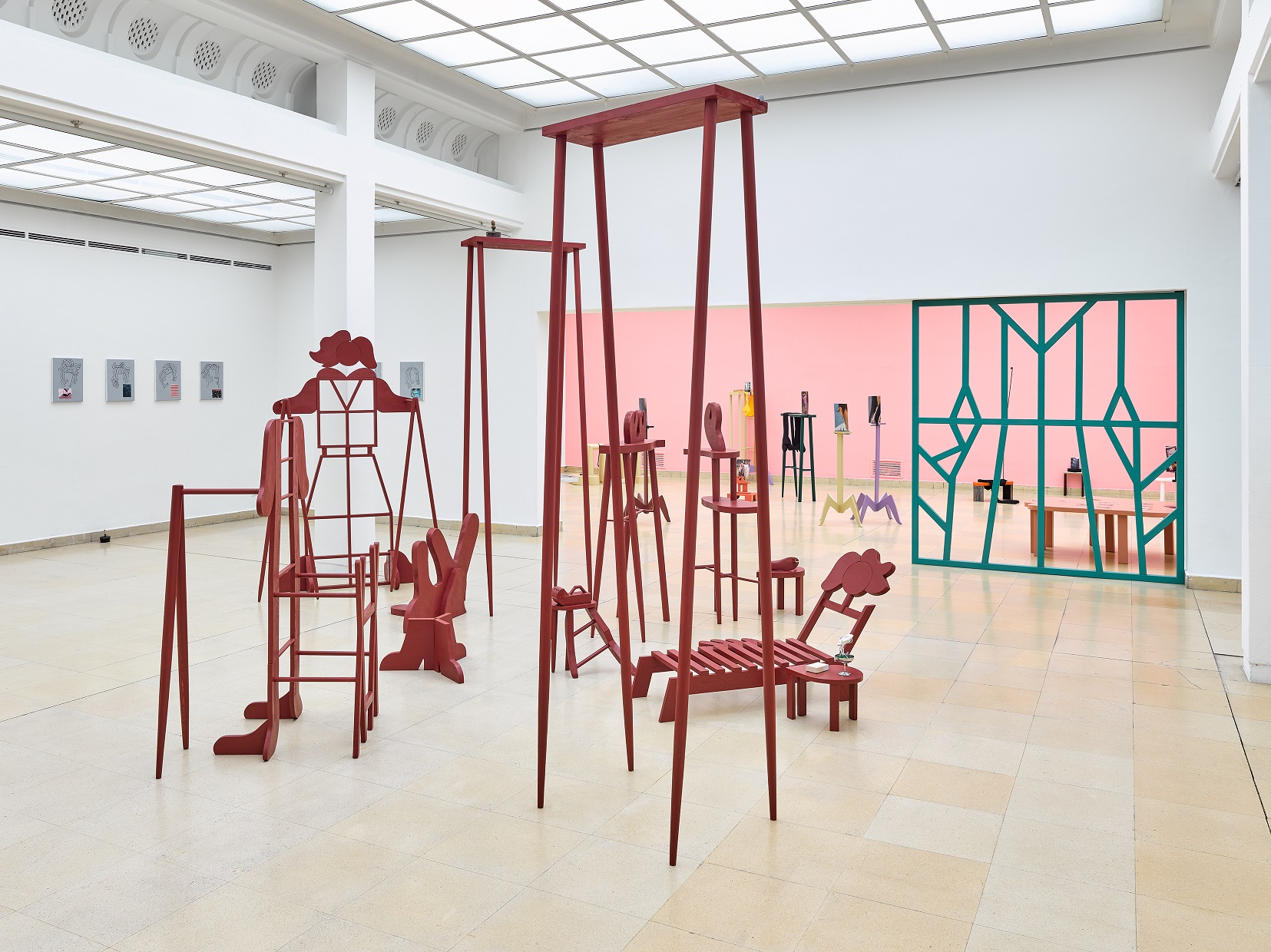 Ausstellungsansicht Kunstpavillon mit rot lackierten Holzskulpturen in unterschiedlichen Größen und Höhen und im Hintergrund ein Raumteiler mit dahinterliegenden Skulpturen vor rosa Wand