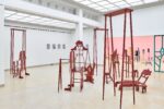 Ausstellungsansicht Kunstpavillon mit rot lackierten Holzskulpturen in unterschiedlichen Größen und Höhen mit Figurenobjekt und links gemalte Bilder