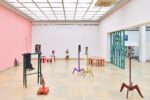 Ausstellungsansicht Kunstpavillon mit unterschiedlich farbig lackierten Holzskulpturen in unterschiedlichen Größen und Höhen mit Fotos vor rosa Wand, rechts ein Wandteiler im Durchgang