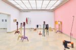 Ausstellungsansicht Kunstpavillon mit unterschiedlich farbig lackierten Holzskulpturen in unterschiedlichen Größen und Höhen mit Fotos vor rosa Wand