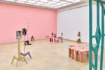 Ausstellungsansicht Kunstpavillon mit unterschiedlich farbig lackierten Holzskulpturen in unterschiedlichen Größen und Höhen mit Fotos vor rosa Wand