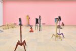 Ausstellungsansicht Kunstpavillon mit unterschiedlich farbig lackierten Holzskulpturen in unterschiedlichen Größen und Höhen mit Fotos vor rosa Wand