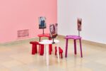 Ausstellungsansicht Kunstpavillon mit unterschiedlich farbig lackierten Holzskulpturen in unterschiedlichen Größen und Höhen mit Fotos vor rosa Wand
