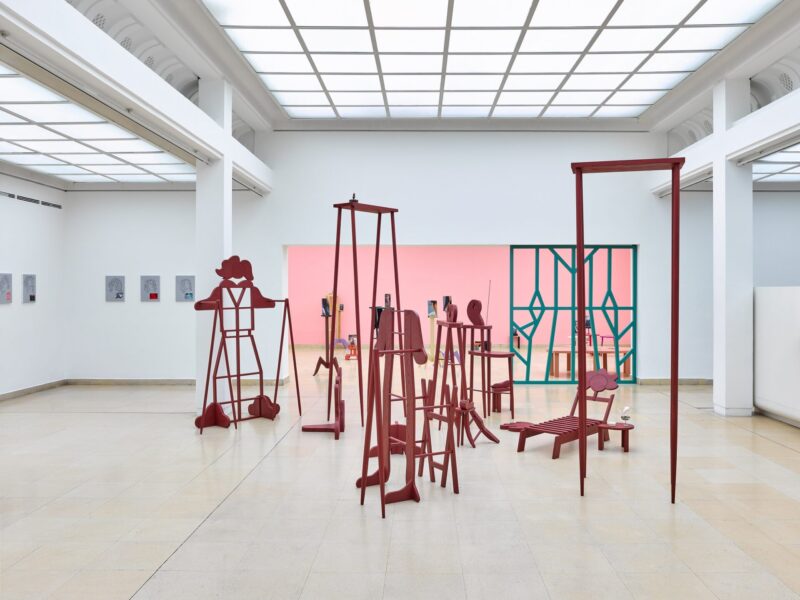 Exhibition view Kunstpavillon with red-painted wooden sculptures in various sizes and heights and pictures on the left and a pink painted back wall