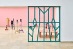 Ausstellungsansicht Kunstpavillon mit Raumteiler und dahinter unterschiedlich farbig lackierten Holzskulpturen in unterschiedlichen Größen und Höhen mit Fotos