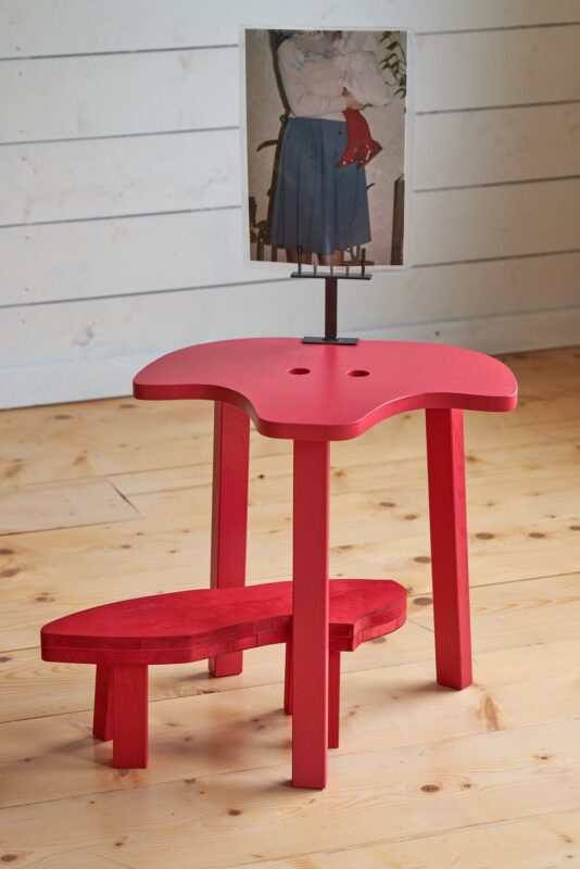 rote höckerähnliche Skulptur mit einem Foto darauf, welches eine Frau zeigt die ein Kind mit roten Lackschuhen trägt, man sieht nur die Oberkörper und Beine der fotografierten Personen; eine weiße Holzwand ist im Hintergrund zu sehen und ein hellbrauner Holzboden