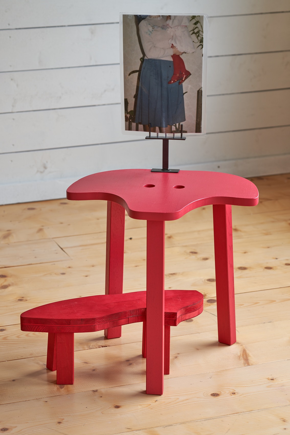 rote höckerähnliche Skulptur mit einem Foto darauf, welches eine Frau zeigt die ein Kind mit roten Lackschuhen trägt, man sieht nur die Oberkörper und Beine der fotografierten Personen; eine weiße Holzwand ist im Hintergrund zu sehen und ein hellbrauner Holzboden