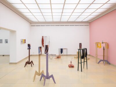 Ausstellungsansicht Kunstpavillon mit unterschiedlich farbig lackierten Holzskulpturen in unterschiedlichen Größen und Höhen mit Fotos