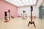 Exhibition view Kunspavillon with wooden sculptures painted in different colours, in various sizes and heights, with photographs next to a pink wall