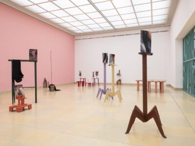Ausstellungsansicht Kunstpavillon mit unterschiedlich farbig lackierten Holzskulpturen in unterschiedlichen Größen und Höhen mit Fotos