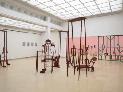 Ausstellungsansicht Kunstpavillon mit rot lackierten Holzskulpturen in unterschiedlichen Größen und Höhen mit Figurenobjekt und links gemalte Bilder