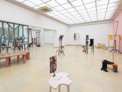Ausstellungsansicht Kunstpavillon mit unterschiedlich farbig lackierten Holzskulpturen in unterschiedlichen Größen und Höhen mit Fotos