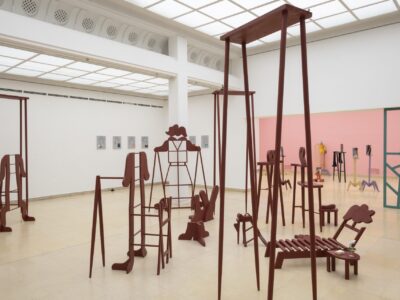 Ausstellungsansicht Kunstpavillon mit rot lackierten Holzskulpturen in unterschiedlichen Größen und Höhen mit Figurenobjekt und links gemalte Bilder