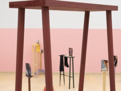 Ausstellungsansicht Kunstpavillon mit kleinem Topf auf einer rot lackierten Holzskulptur mit bunten Skulpturen und Fotos im Hintergrund