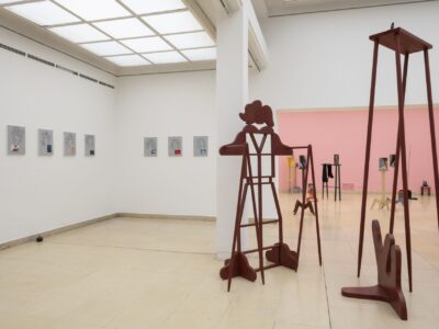 Ausstellungsansicht Kunstpavillon mit rot lackierten Holzskulpturen in unterschiedlichen Größen und Höhen mit Figurenobjekt und links gemalte Bilder