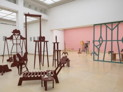 Ausstellungsansicht Kunstpavillon mit unterschiedlich farbig lackierten Holzskulpturen in unterschiedlichen Größen und Höhen mit Fotos, links gemalte Bilder und im Hintergrund Raumteiler aus Holz