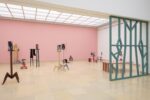 Exhibition view Kunspavillon with wooden sculptures painted in different colours, in various sizes and heights, with photographs in front to a pink wall