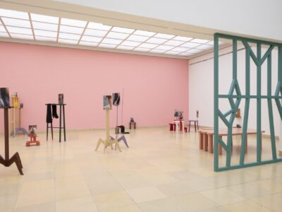 Ausstellungsansicht Kunstpavillon mit unterschiedlich farbig lackierten Holzskulpturen in unterschiedlichen Größen und Höhen mit Fotos vor rosa Wand