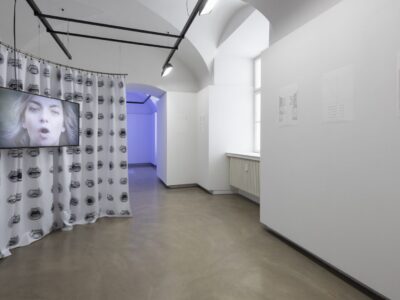 Ausstellungsansicht, im vorderen Raum sieht man einen Vorhang der in einer runden Form hängt mit Mündern darauf und ein Screen mit einem Gesicht, im hinteren Raum scheint blaues Licht