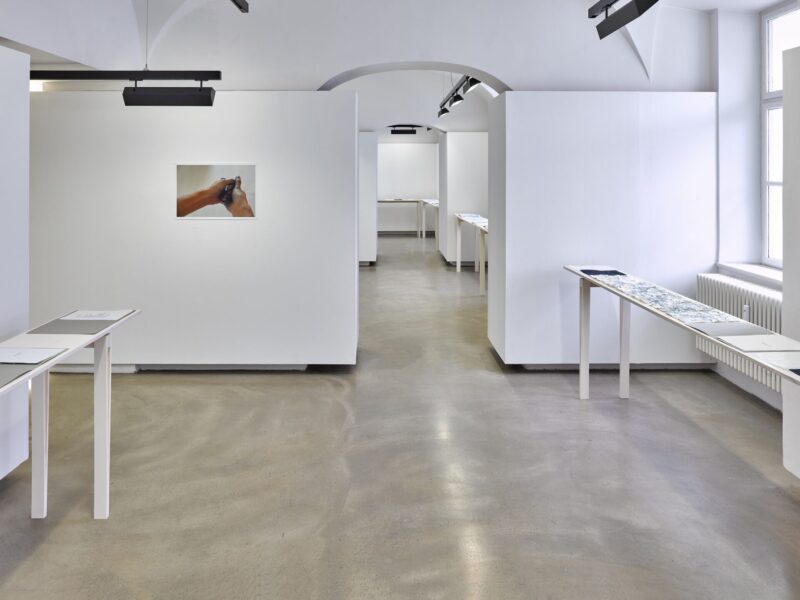 Foto Ausstellung Neue Galerie mit schmalen weiß lasierten Holztischen und einem blauen Papierblätt darauf, daneben eine augeschlagene graue Mappe mit einem mit Scheibmaschine beschriebenem Blatt, an der Wand ein Foto einer Hand, die ein dunkles Blatt zerdrückt