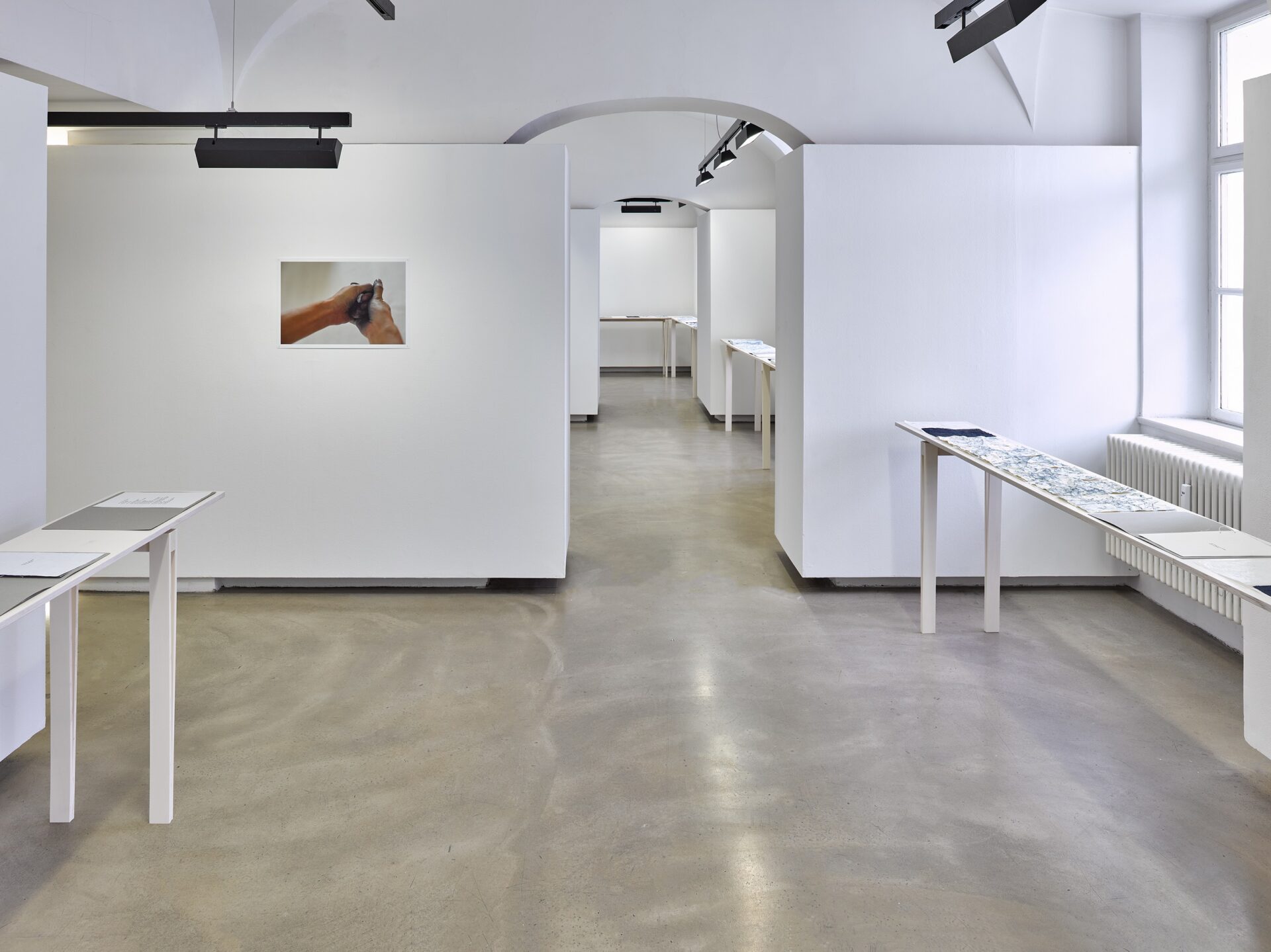 Photo exhibition Neue Galerie with narrow white stained wooden tables and blue sheets of paper on top, next to it an open grey folder with a sheet of paper written on with a typewriter, on the wall a photo of a hand crushing a dark sheet of paper.