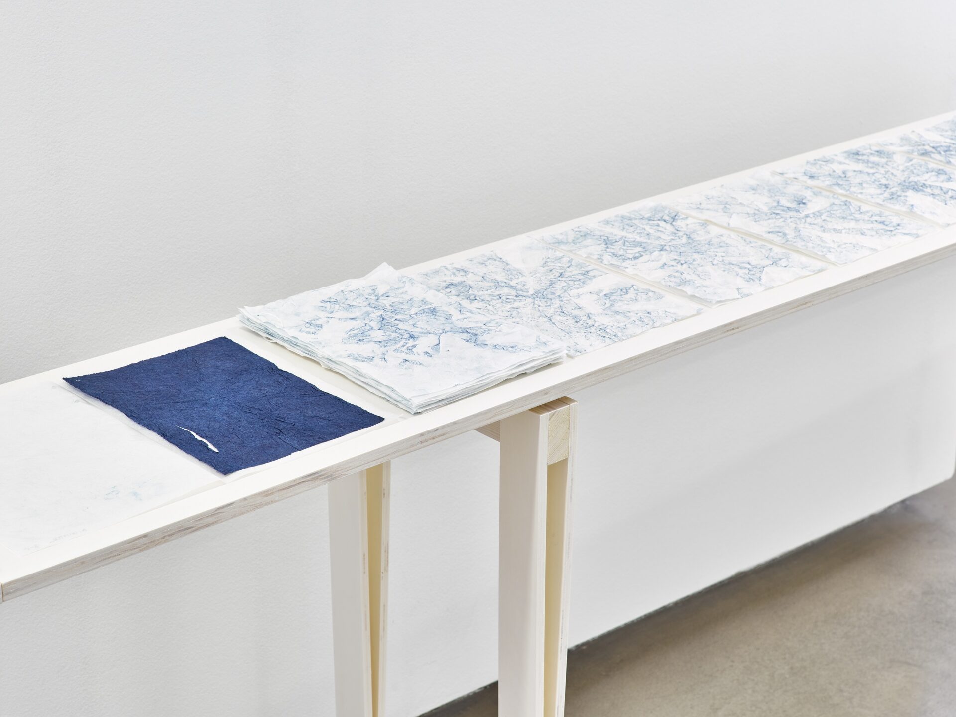 Photo exhibition Neue Galerie with narrow white-stained wooden tables and blue and blue-white sheets of paper on them