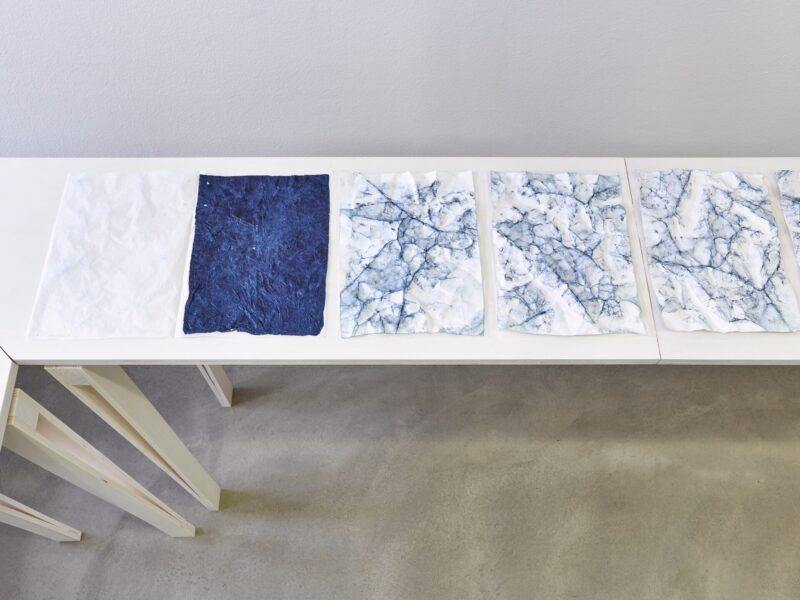 Foto Ausstellung Neue Galerie mit schmalen weiß lasierten Holztischen und weißblaue, ein blaues und ein weißes blauen Papierblätter darauf