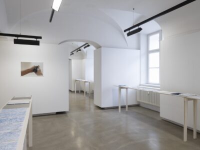 Foto Ausstellung Neue Galerie mit schmalen Tischen und weißen, blauweißen und blauen Blättern darauf, an der Wand ein Foto von einer Hand die etwas dunkles zerdrückt