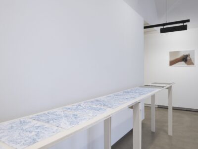 Foto Ausstellung Neue Galerie mit schmalen Tischen mit darauf gelegten blauweißen und weißen Papierblättern und grauen aufgeschlagenen Aktenmappe, an der Wand ein Foto von einer Hand die etwas dunkles zerdrückt