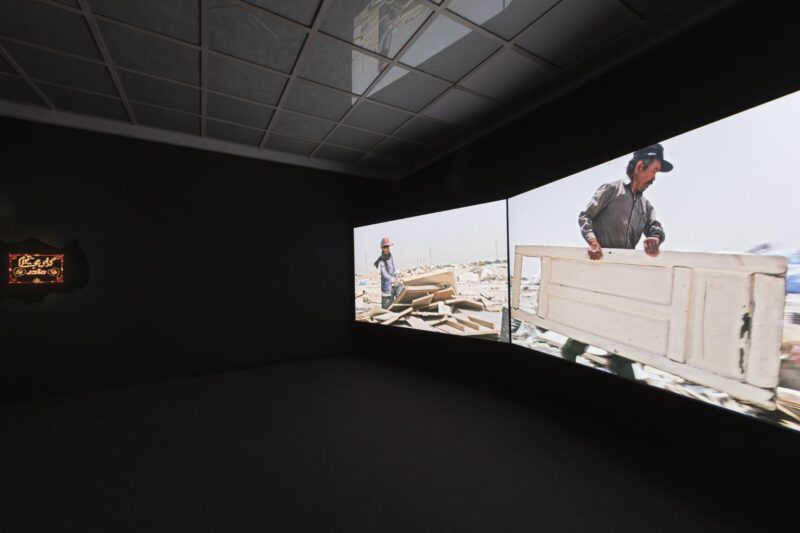 Foto aus Ausstellung zwei große projizierte Bilder in einem schwarzen Raum und ein Bild mit rot und weißer Schrift