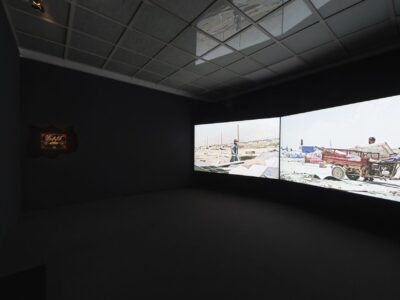 Foto aus Ausstellung zwei große projizierte Bilder in einem schwarzen Raum und ein Bild mit rot und weißer Schrift