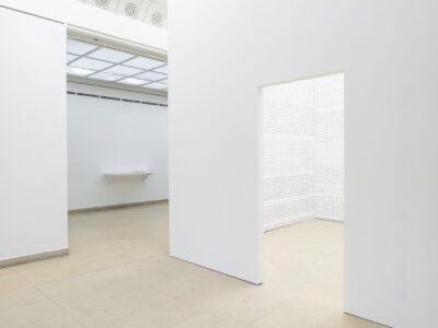 Foto aus Ausstellung mit weißen Raum im Ausstellungsraum mit Reihen im Hintergrund ein Regal