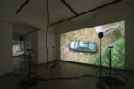 Photo exhibition with videos and text on the wall, speakers and small projectors on tripods on the floor