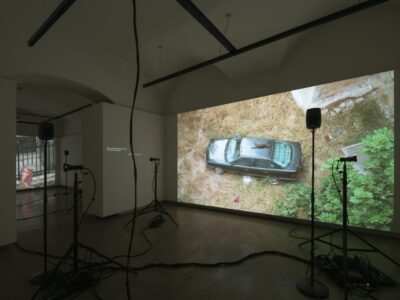 Foto Ausstellung mit Videos und Text an den Wänden, Lautsprecher und kleine Projektoren auf Stativen stehen am Boden